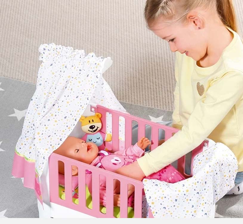 Mädchen spielt mit Puppe in rosa Gitterbett mit Spielzeugvorhang und Teddy.