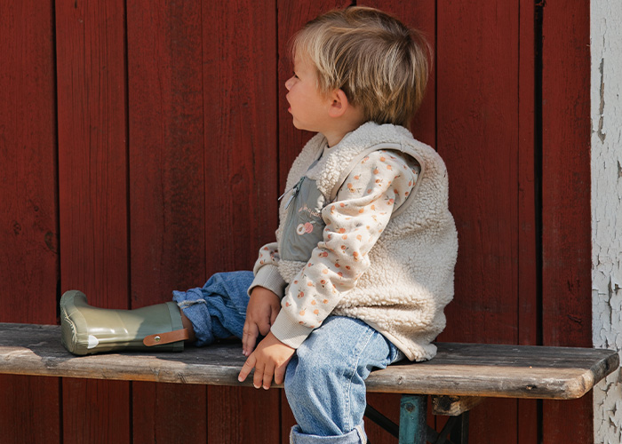 Ein Kind sitzt auf einer Bank in einer Weste und Gummistiefeln, mit dem Text 'Kenza X Viking'.