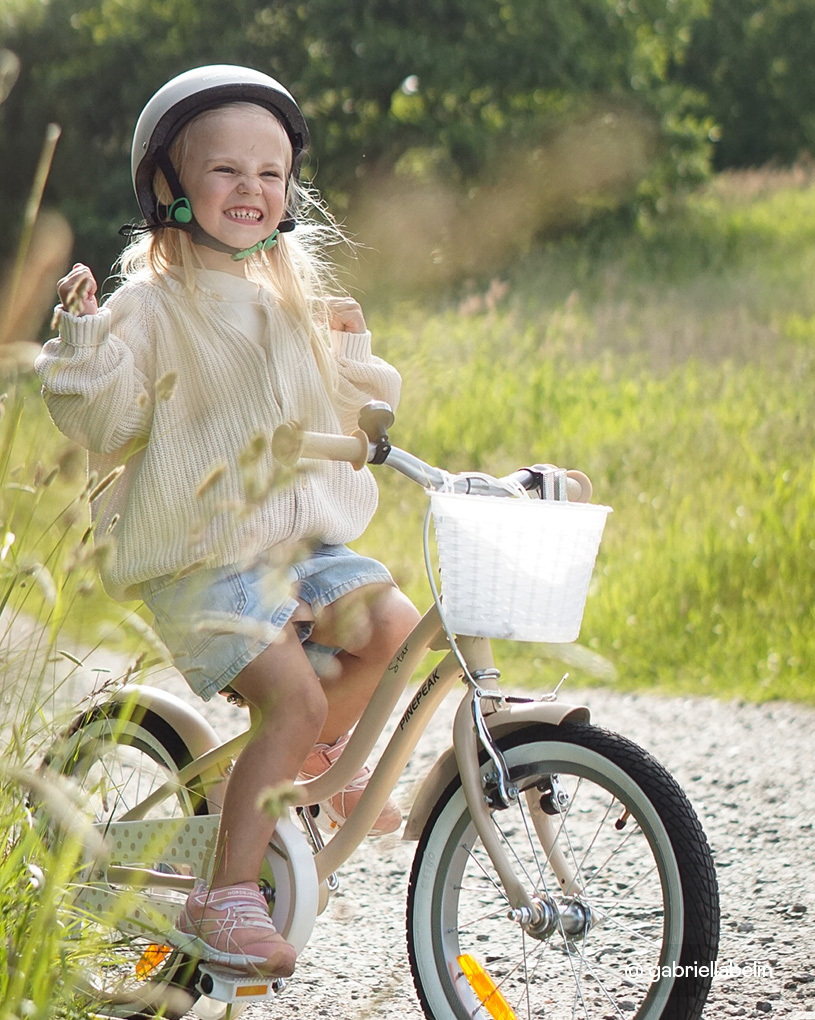 Mädchen mit Helm fährt ein beiges Pinepeak-Fahrrad mit weißem Korb auf einem Kiesweg in der Sonne.
