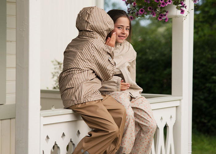 Zwei Kinder sitzen auf einem Balkon und flüstern sich etwas zu, mit dem Text 'Kenza X Viking'.