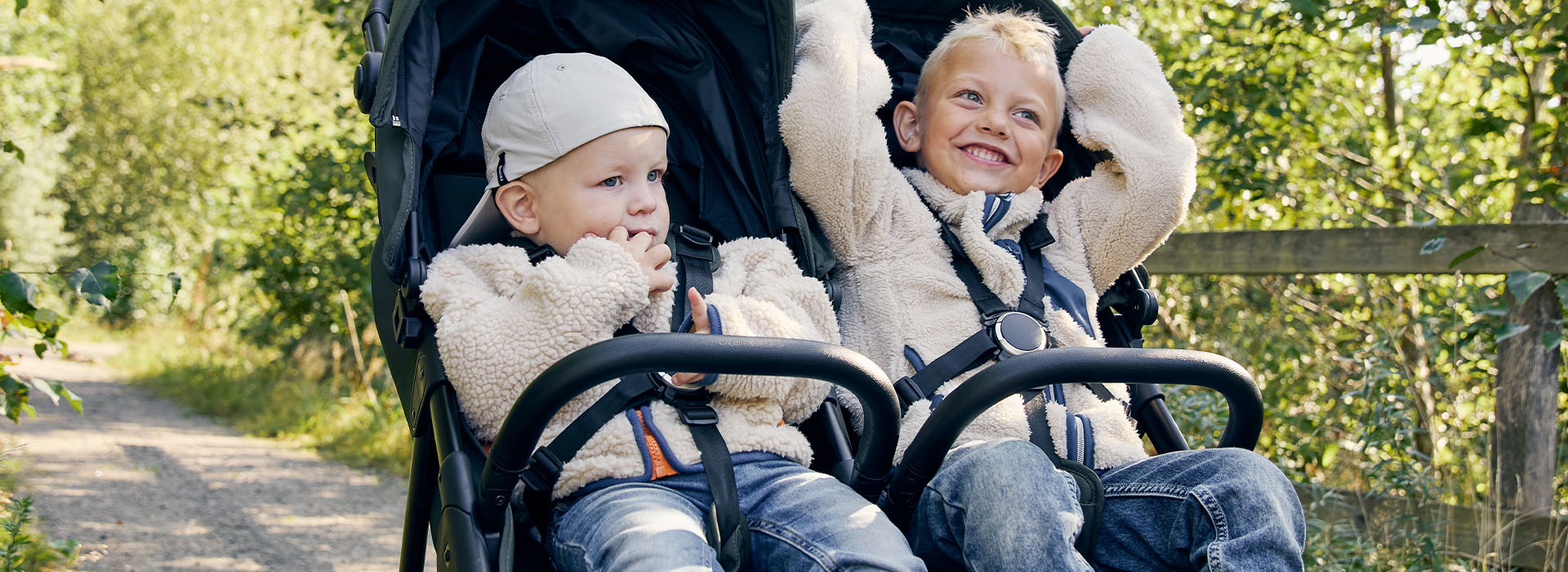 Zwei lächelnde Kinder im Kinderwagen auf Waldweg.
