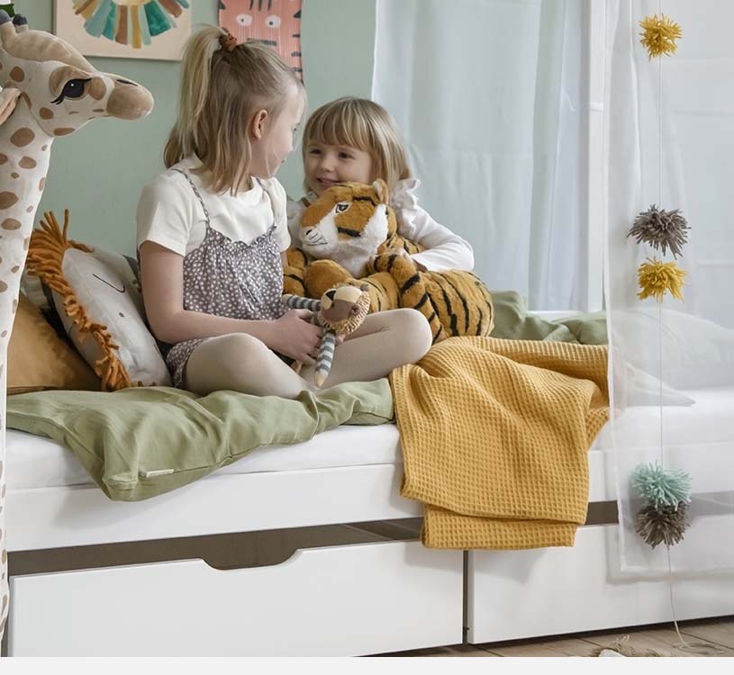 Zwei Kinder spielen mit Plüschtieren, Giraffe und Tiger, auf dem Bett.